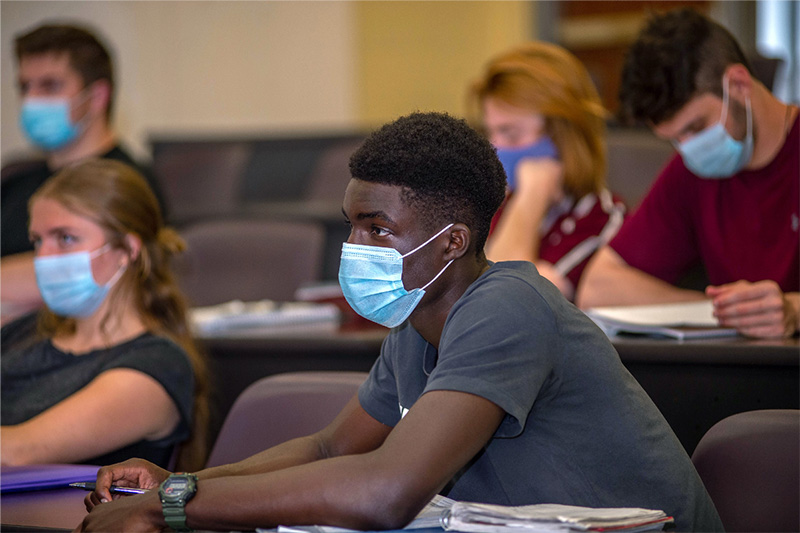 Students In Masks In A Classroom Listening To A Lecture.