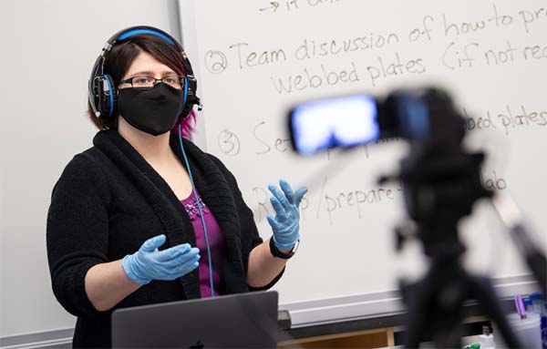 Faculty member in mask and protective gloves teaching in front of a classroom.