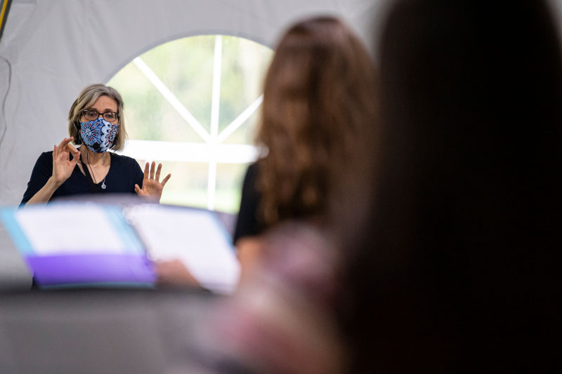 Faculty member addressing students in her class from front of room while wearing a mask.